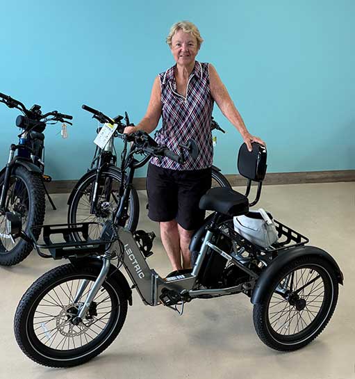 Woman-Lectric-Trike-Ocala An older woman stands indoors, smiling and holding the handlebars of an electric tricycle. She wears a sleeveless patterned shirt and black shorts. Several other bikes are visible in the background against a blue wall.