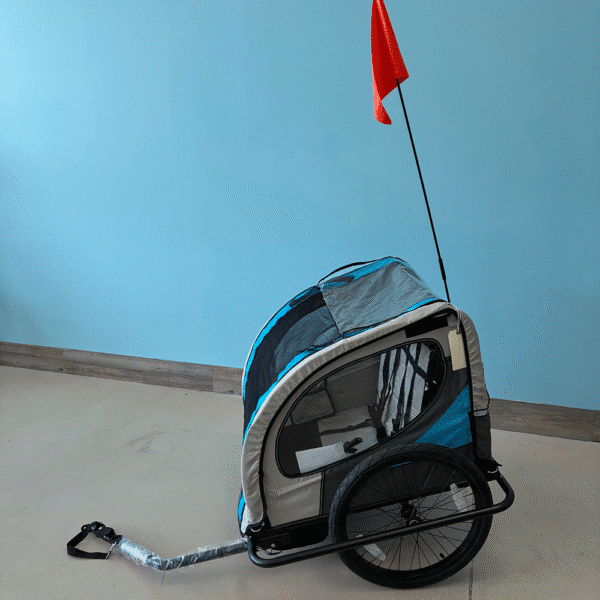 A blue and gray Kids eBike Trailer with two wheels and a transparent cover sits indoors against a light blue wall. A red safety flag is attached to a tall pole on the trailer.