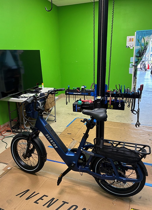 Bike Repair Area A blue Aventon electric bike is mounted on a repair stand in a workshop with a bright green wall, tools hanging in the background, and a TV on a nearby table.