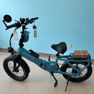 A blue Lectric folding electric bike is parked indoors against a light blue wall. The bike has a rear rack holding a small box and various accessories attached to the handlebars.