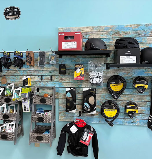 carousel-ebikes-shop-wall A display wall in a bike shop shows helmets, gloves, bike pedals, cycling accessories, and gear hanging neatly on rustic wooden shelves against a light blue wall.