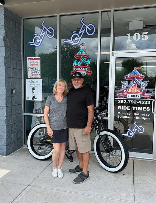 dawn-rj-bike-shop-ocala A smiling couple stands in front of a shop with glass doors, a black bicycle with white tires, and bright bike-themed signs. The woman wears a gray shirt and shorts; the man wears a black shirt, shorts, and a red cap.