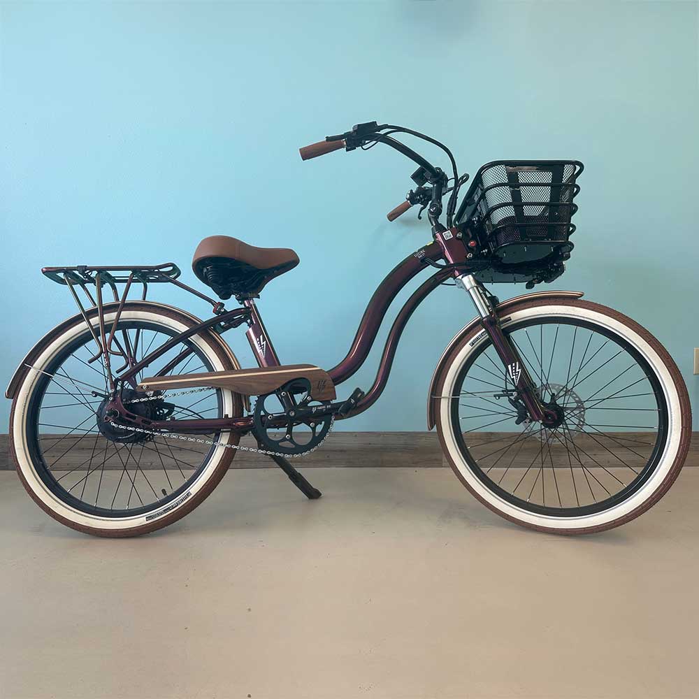 A maroon cruiser bicycle with brown seat and handlebar grips, whitewall tires, a front basket, and a rear cargo rack, standing indoors against a light blue wall.
