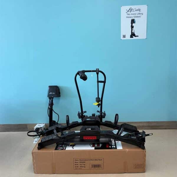 A black Hollywood E-Bike Rack and Lift Caddy for Cars is displayed on a cardboard box against a light blue wall. A small poster reads, "No more lifting heavy e-bikes." To the left are a power outlet and a black device.