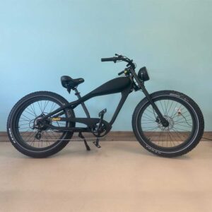 A matte black Revi Black Cheetah Plus eBike with fat tires, upright handlebars, and a padded seat is parked indoors against a light blue wall and beige floor.