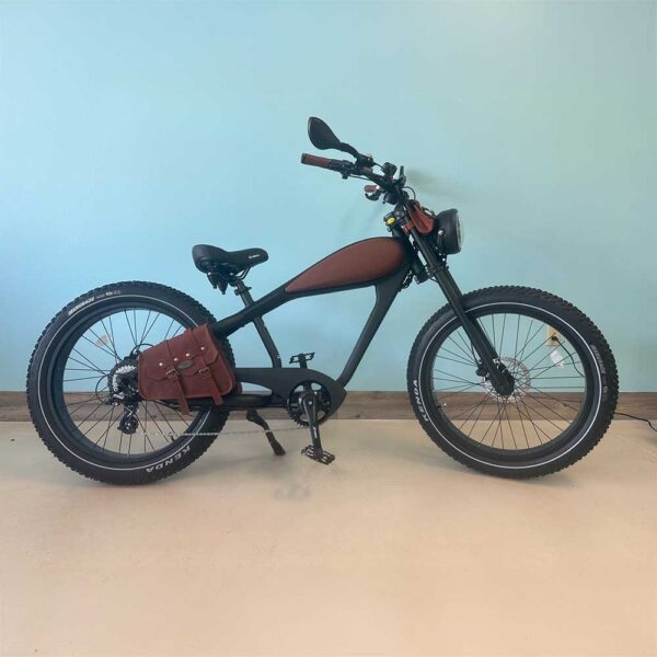 A Revi Brown Cheetah Plus eBike with accessories, including a brown leather saddle, matching side bag, and wide tires, is parked indoors against a pale blue wall.