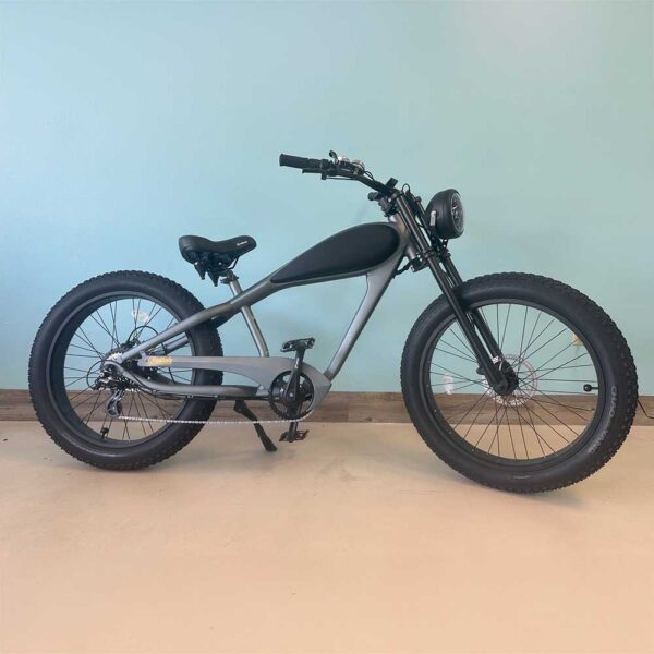A matte black and gray Revi Silver & Black Cheetah eBike cruiser with large, thick tires is parked indoors against a light blue wall and tan floor.