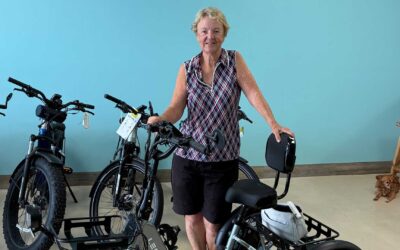 A smiling older woman stands indoors, holding onto an electric bike. Several other bikes are nearby. She wears a sleeveless patterned shirt and shorts. A small brown dog sits on the floor in the background.