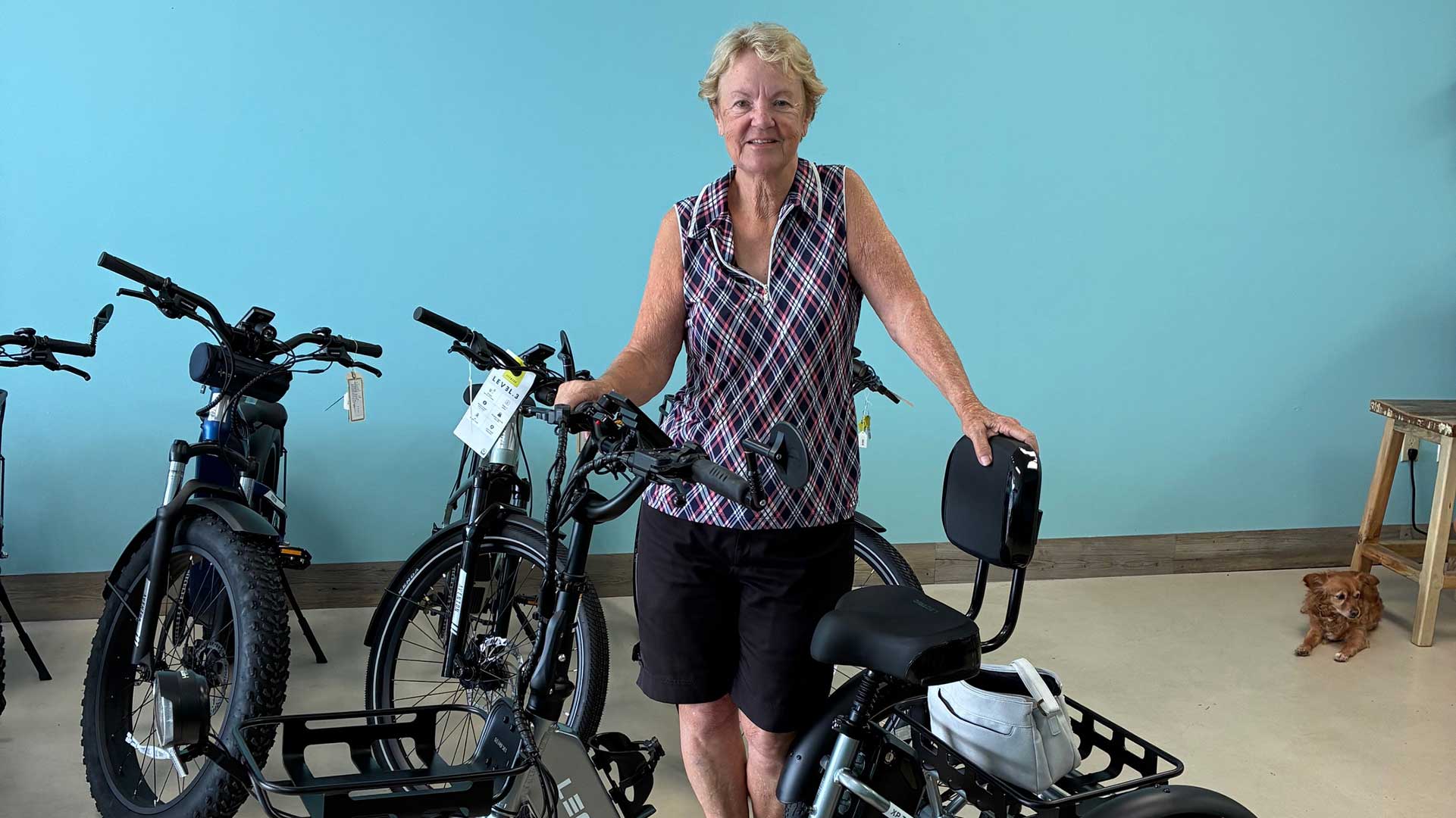 A smiling older woman stands indoors, holding onto an electric bike. Several other bikes are nearby. She wears a sleeveless patterned shirt and shorts. A small brown dog sits on the floor in the background.