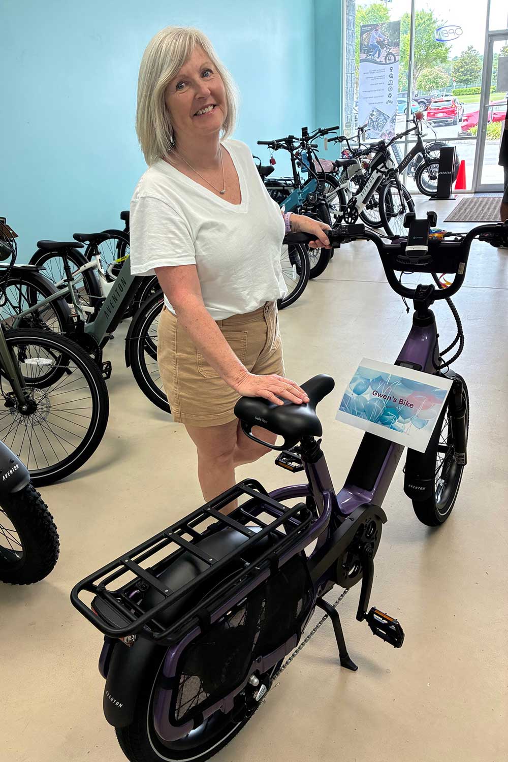 A smiling woman with gray hair, wearing a white t-shirt and beige shorts, stands indoors beside a purple electric bike in a bike shop. Several other bikes are displayed in the background.