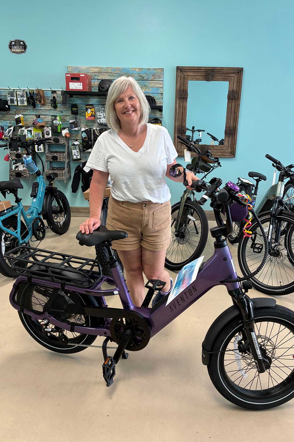 A smiling woman with gray hair, wearing a white t-shirt and tan shorts, stands indoors holding a purple Aventon electric bike in a bike shop with other bikes and accessories on display.