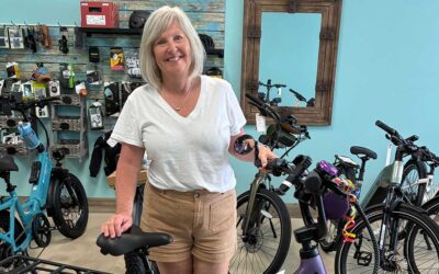 A smiling woman with gray hair, wearing a white t-shirt and beige shorts, stands in a bike shop holding the handlebars of a bicycle. Several other bikes and accessories are visible in the background.