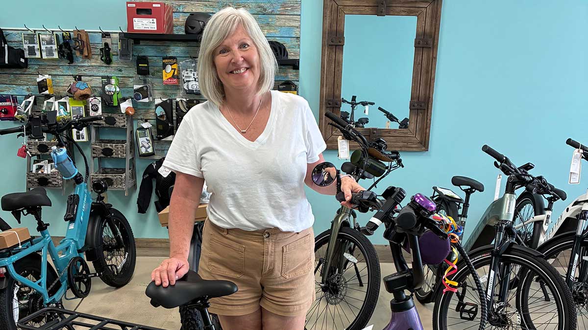 A smiling woman with gray hair, wearing a white t-shirt and beige shorts, stands in a bike shop holding the handlebars of a bicycle. Several other bikes and accessories are visible in the background.