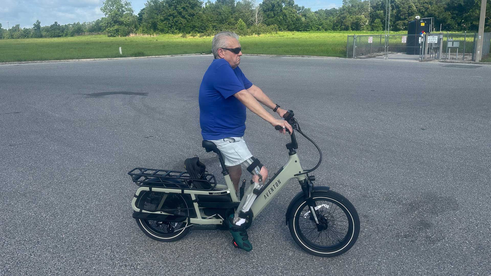 An older man wearing sunglasses, a blue shirt, and white shorts is riding a light-colored electric bike in an empty paved lot with grass and trees in the background.