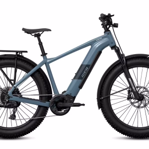A matte blue electric fat-tire bike with a sturdy frame, front suspension, rear cargo rack, disc brakes, and thick black tires, shown in a side profile on a white background.
